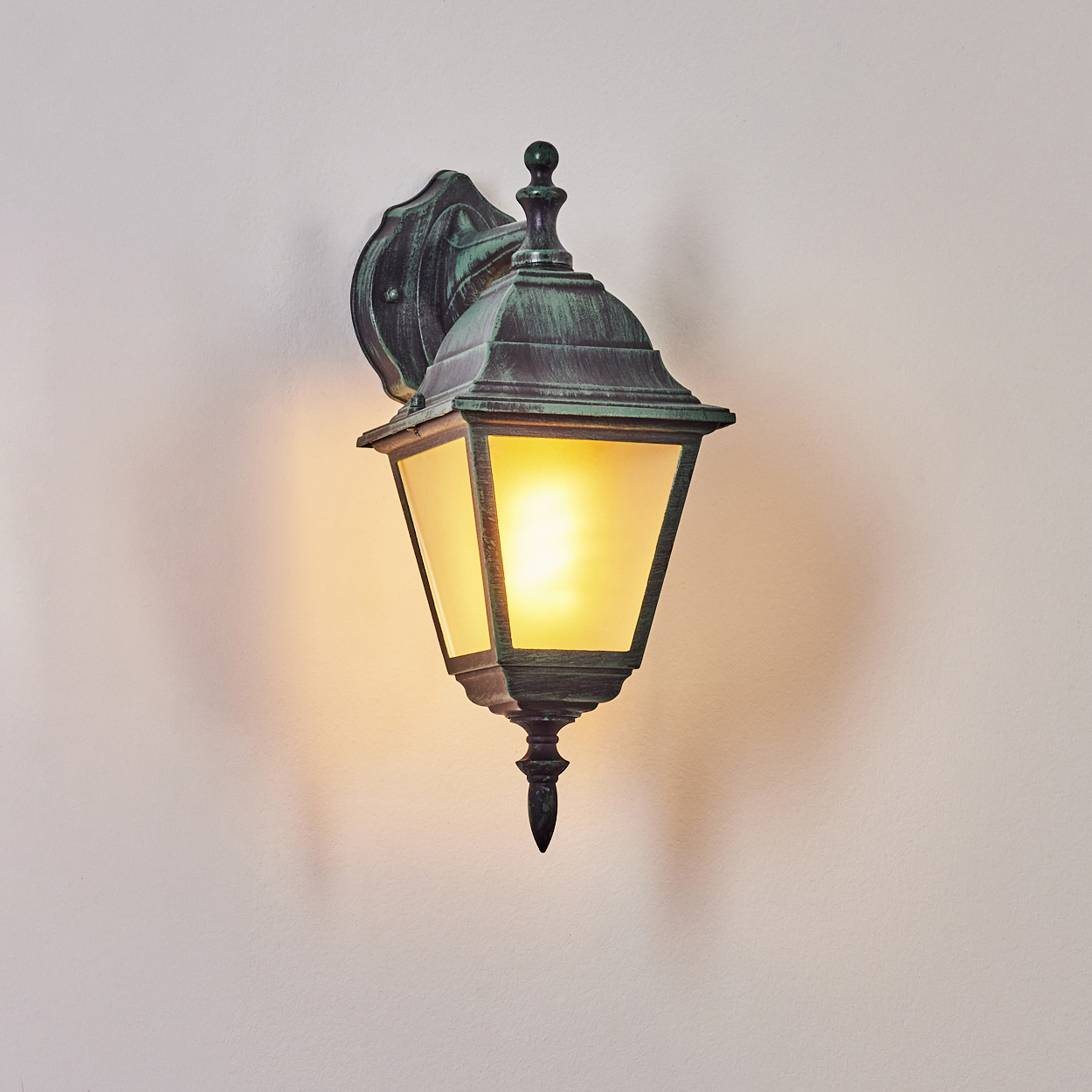 Eine Außenlampe mit grünem Metallrahmen, Milchglas und einer beleuchteten Glühbirne, vor einer weißen Wand.