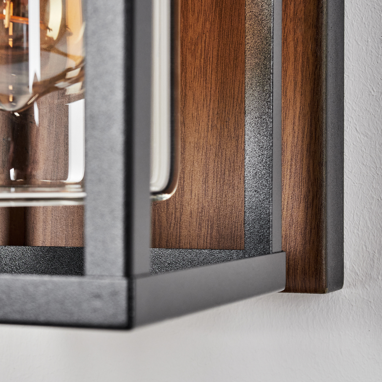 Eine Nahaufnahme einer Wandleuchte mit Holzrücken, schwarzem Rahmen und einem Glasgehäuse um eine Glühbirne. Die Holzmaserung ist sichtbar.