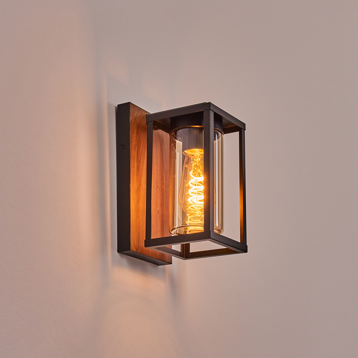Eine Wandlampe mit Holzrückseite, schwarzem Rahmen, Glaszylinder und einer warmen Glühbirne darin.