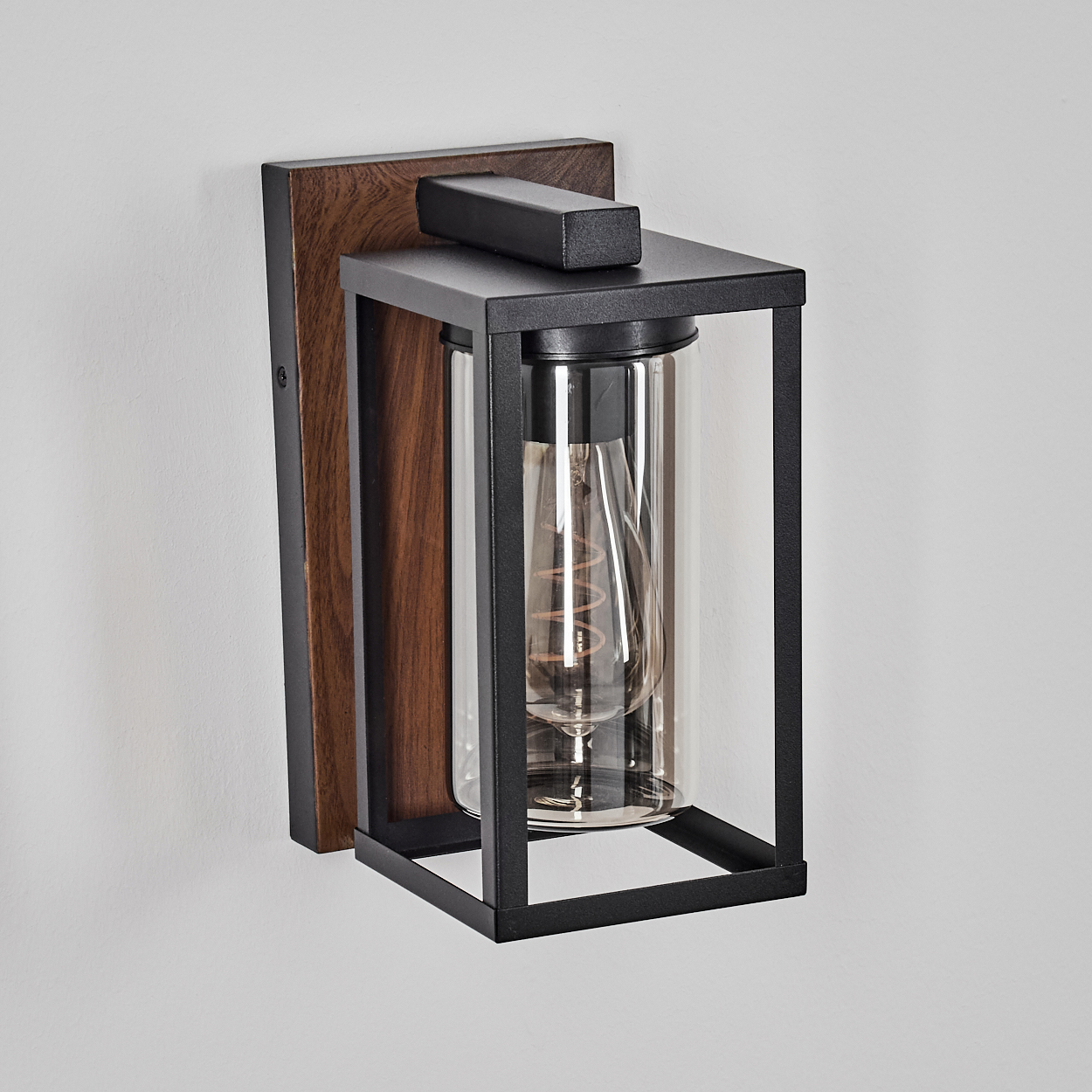Eine Wandlampe mit Holzrücken, schwarzem Rahmen, Glaszylinder und einer warmen Glühbirne im Inneren.