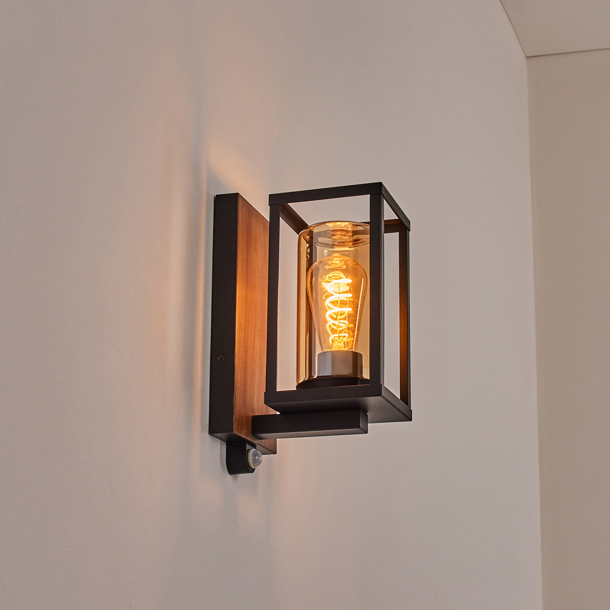 Eine Wandlampe mit Holzsockel, schwarzem Rahmen, Glaszylinder und eingeschalteter Glühbirne.
