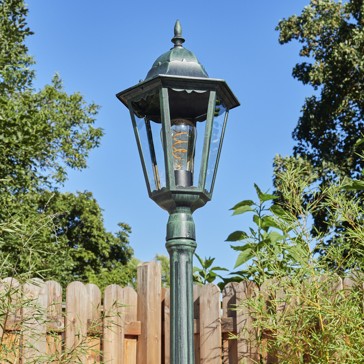 Außenlampe mit grünem Finish. Sie wird von blauem Himmel, Bäumen und einem Holzzaun umrahmt.