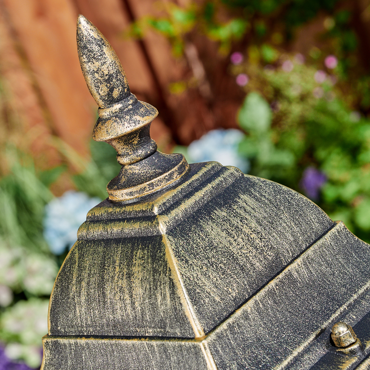 Nahaufnahme eines dekorativen, verzierten Lampenaufsatzes mit Gold- und Schwarzfinish, vor verschwommenem Garten.