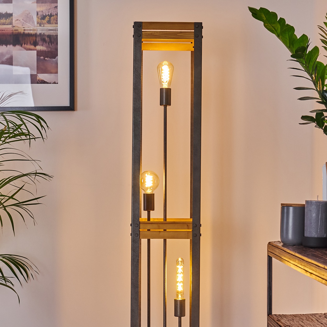 Hohe hölzerne Stehlampe mit drei freiliegenden Glühbirnen. Sie befindet sich in einem Raum mit einem Gemälde und Pflanzen.
