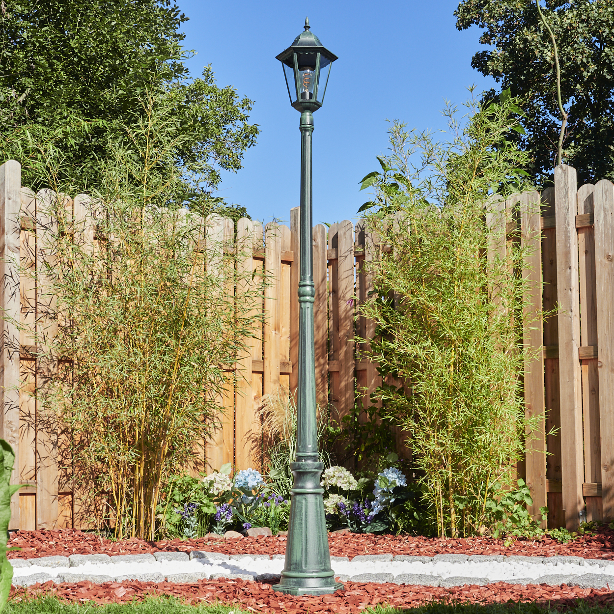 Ein hoher, grüner Außenmast steht in einem Garten mit Holzzaun und Pflanzen.