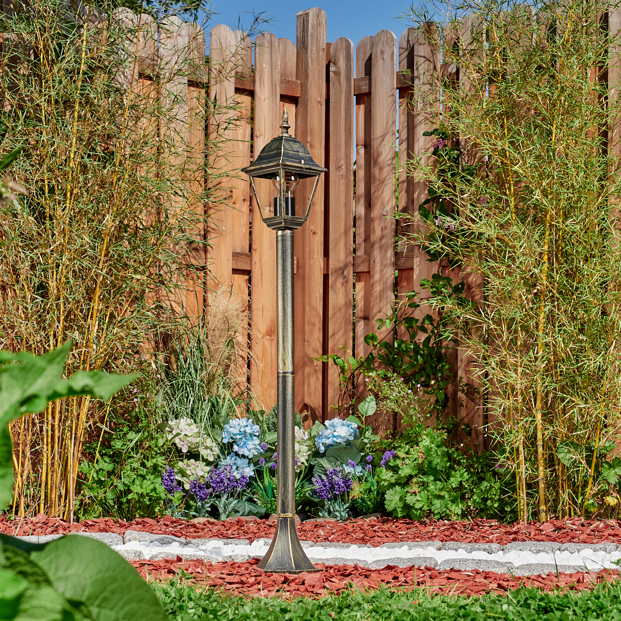 Eine Außenlampe mit goldenem Finish, in einem Garten mit Bambuspflanzen und Holzzaun im Hintergrund.