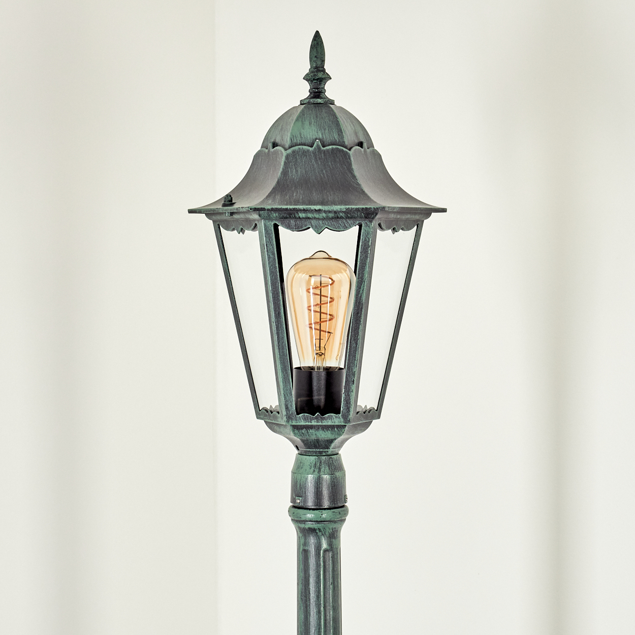 Eine grüne Außenlampe mit klarer Glasabdeckung und einer Wendelglühbirne. Der Hintergrund ist eine weiße Wand.