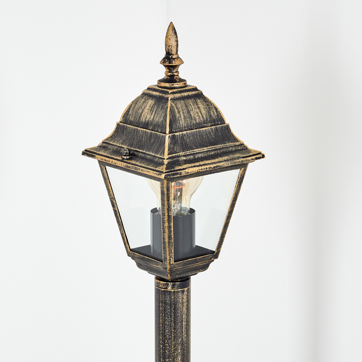 Eine goldene und schwarze Außenlampe mit klarer Glasabdeckung und einer Glühbirne. Der Hintergrund ist eine weiße Wand.