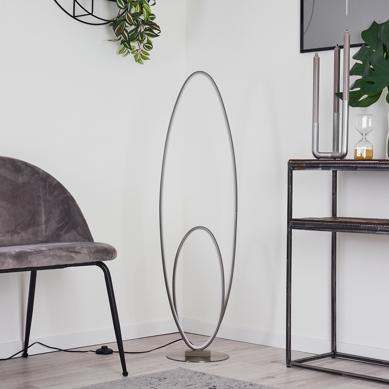 Eine hohe, ovale Stehleuchte in einem Raum mit Stuhl, Tisch und Pflanzen. Die Lampe ist an.