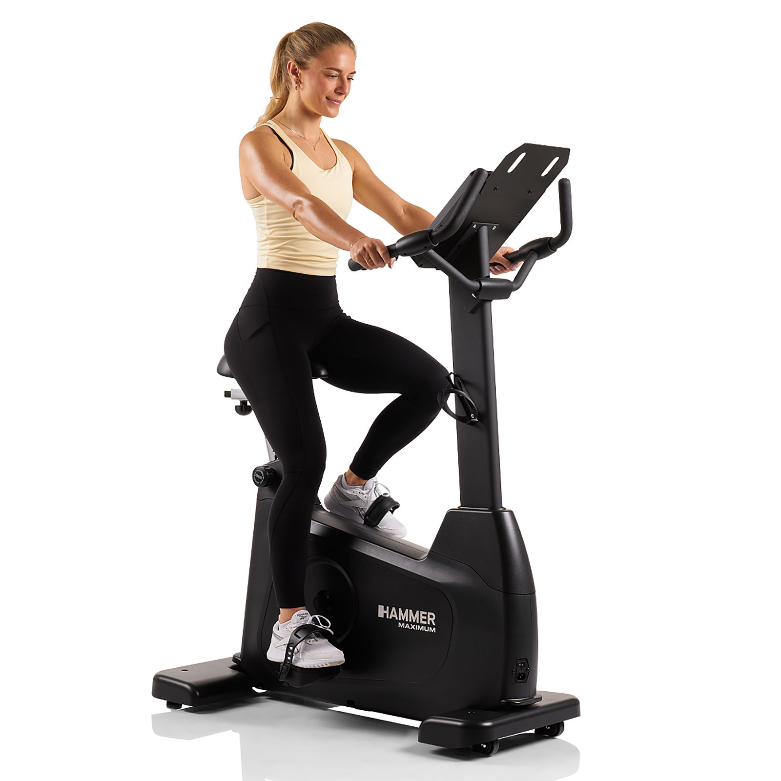 Eine Frau trainiert lächelnd auf einem schwarzen Heimtrainer, trägt schwarze Leggings und ein gelbes Oberteil.