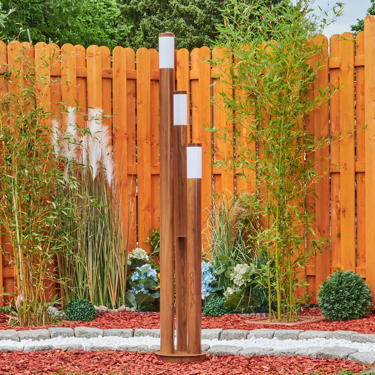 Eine Holzlampe mit drei Holzpfählen und weißen Lichtern in einem Garten mit Holzzaun.