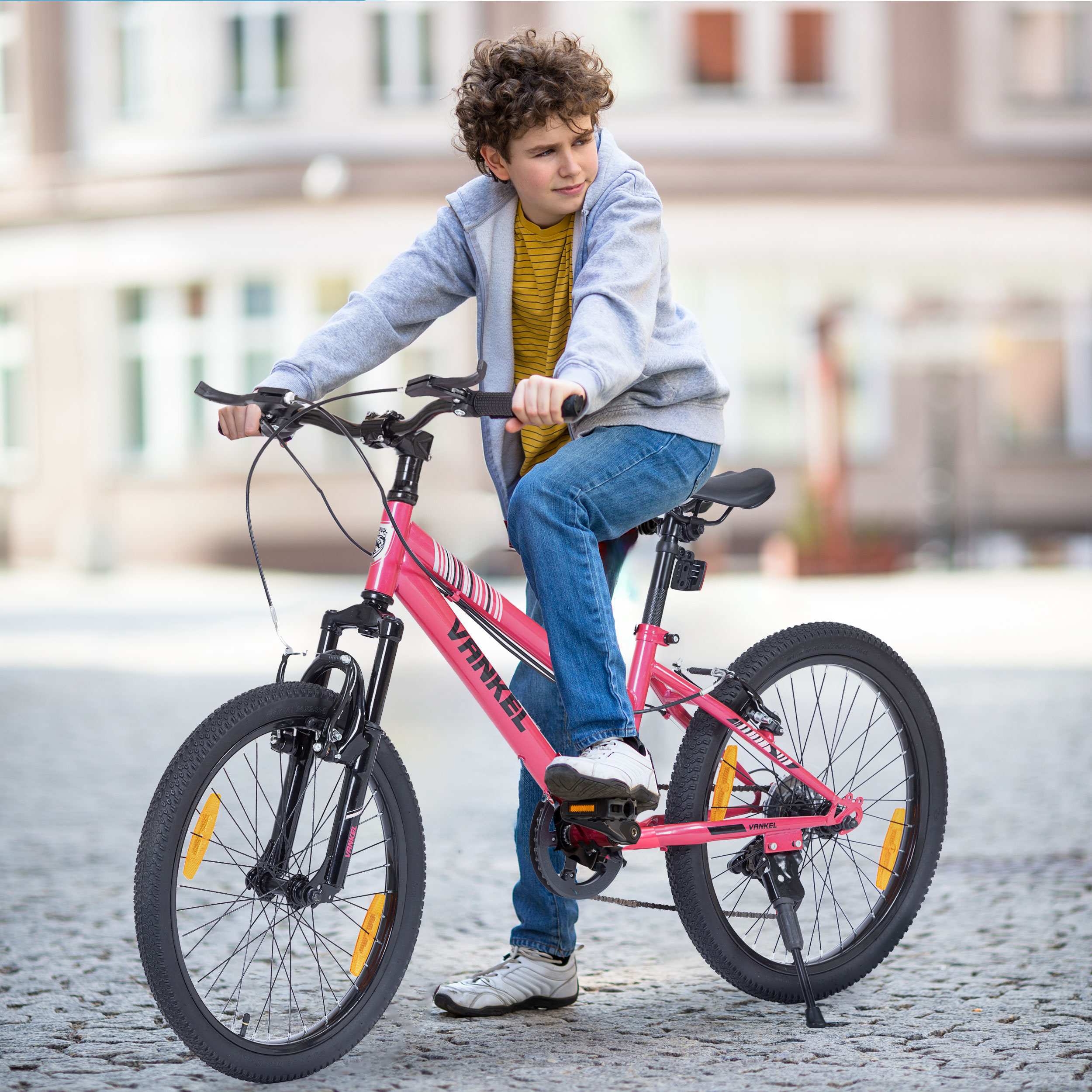 Ein Junge sitzt auf einem rosa Fahrrad auf einer asphaltierten Straße. Er trägt eine graue Jacke, ein gelbes Hemd und blaue Jeans. Gebäude sind im Hintergrund.