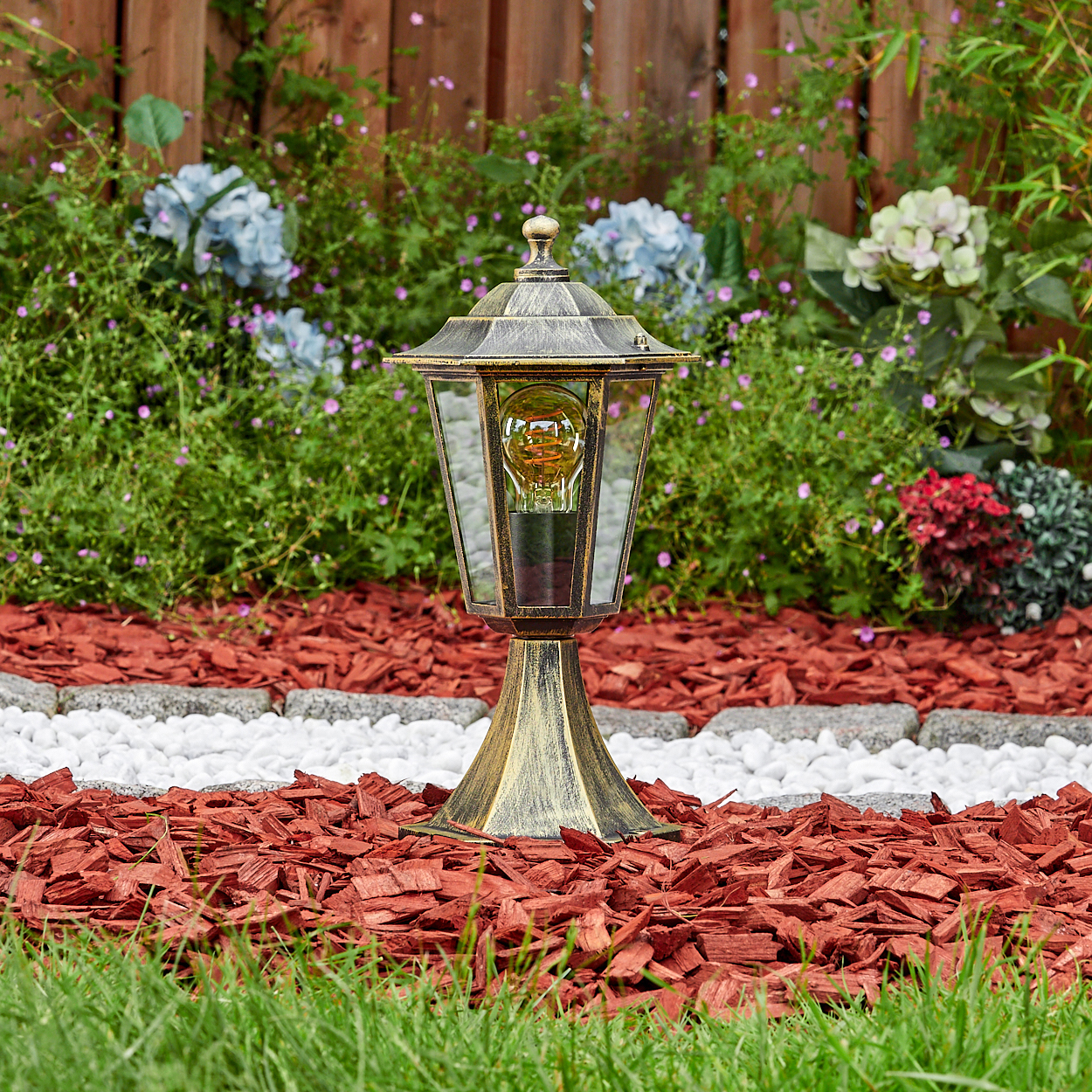 Eine bronzefarbene Lampe mit Glaspaneel und Glühbirne steht auf roter Mulch und Gras, umgeben von grünen Pflanzen.