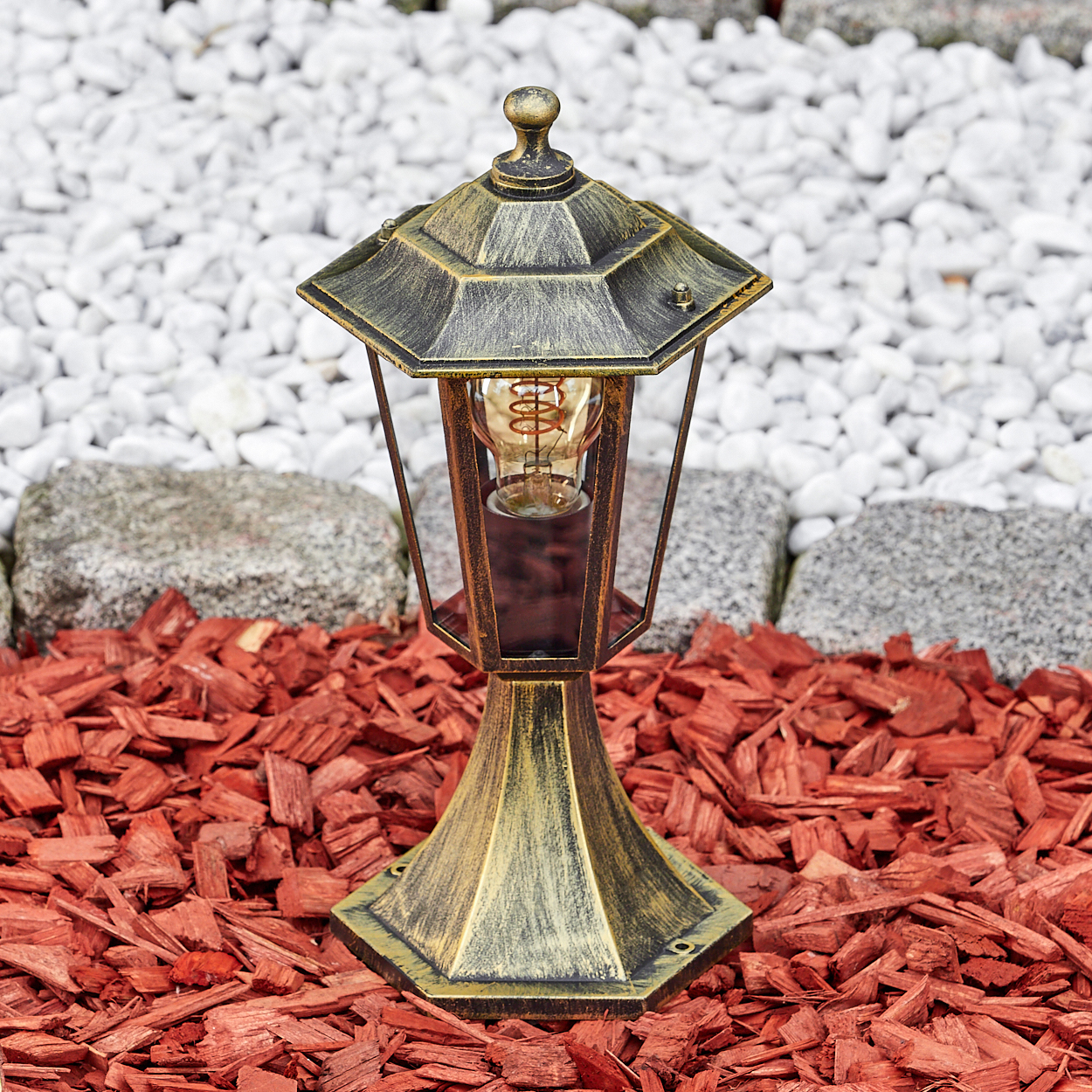 Eine goldene Straßenlaterne mit einem einzelnen Licht, auf rotem Mulch und weißen Steinen in einem Garten.