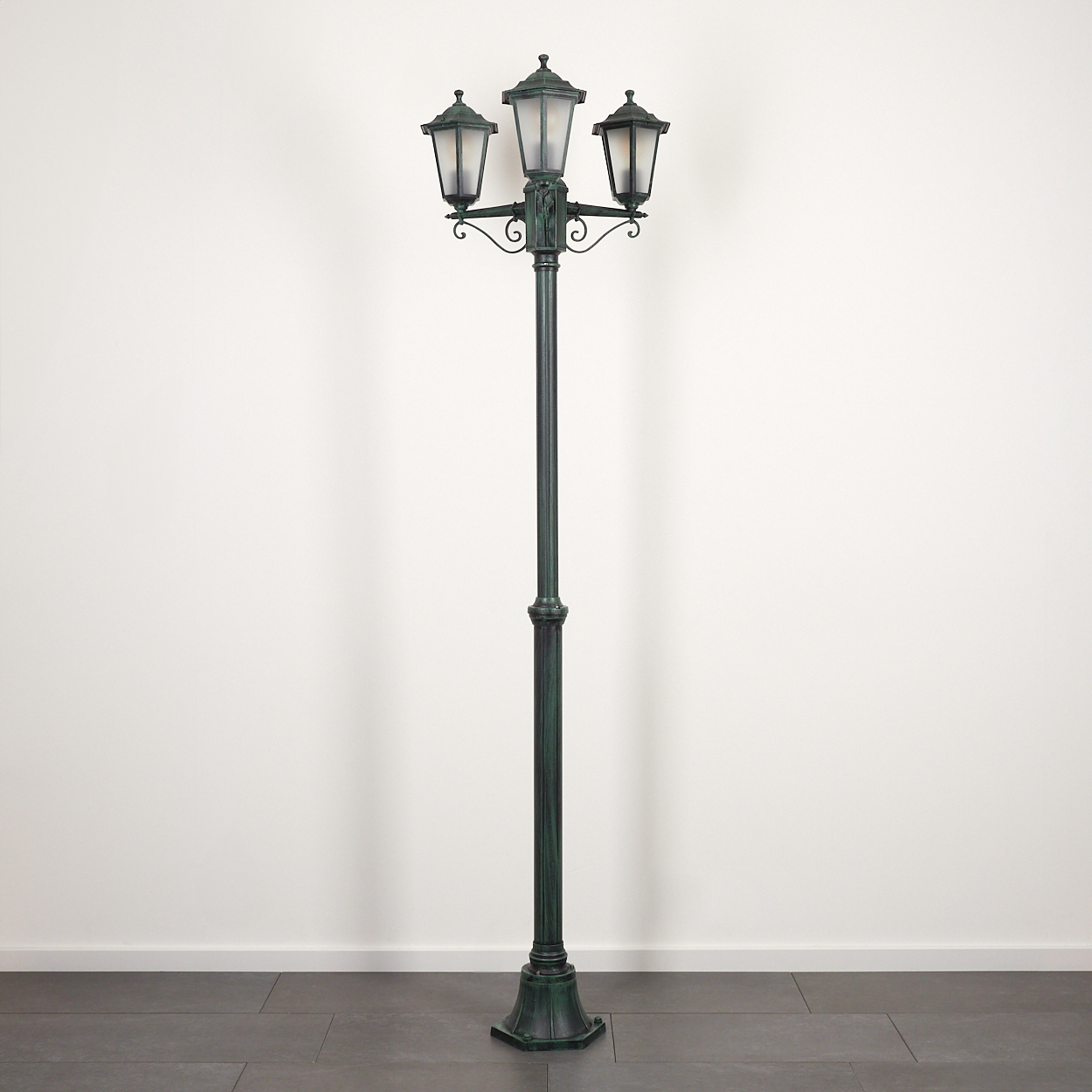 Eine hohe grüne Straßenlaterne mit drei Lampen. Sie steht vor einer weißen Wand und dunklem Boden.