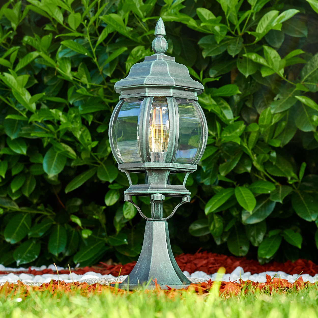 Eine Außenlampe mit grüner Oberfläche steht in einem Garten mit grünen Pflanzen und roter Mulch.
