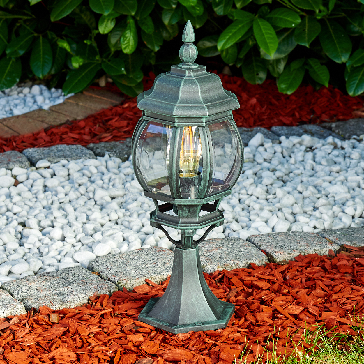 Eine grüne Lampe mit klarem Glas, in einem Garten, auf roter Mulch und weißen Steinen.