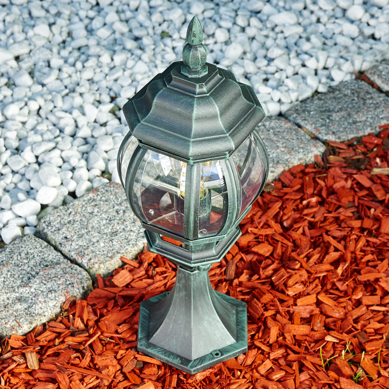 Eine Außenlampe mit grünem Dach und Sockel, auf roter Mulch, mit weißen Steinen und grünem Gras.