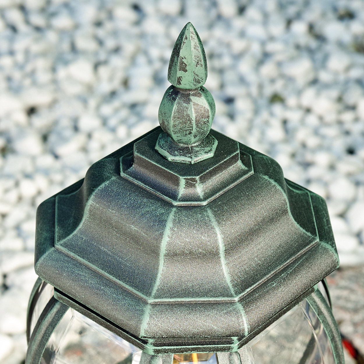 Nahaufnahme der Oberseite einer grünen Außenlampe mit dekorativem Aufsatz auf weißen Steinen.