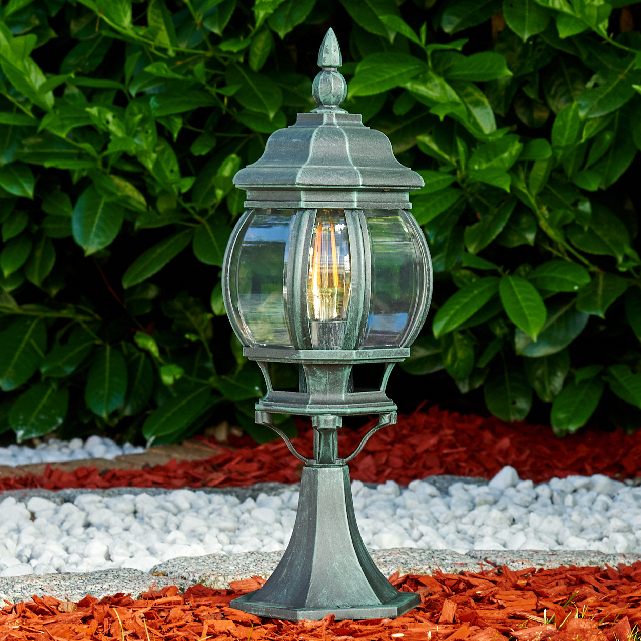 Eine grüne Lampe in einem Garten. Sie hat eine Glaskugel und steht auf roter Mulch.