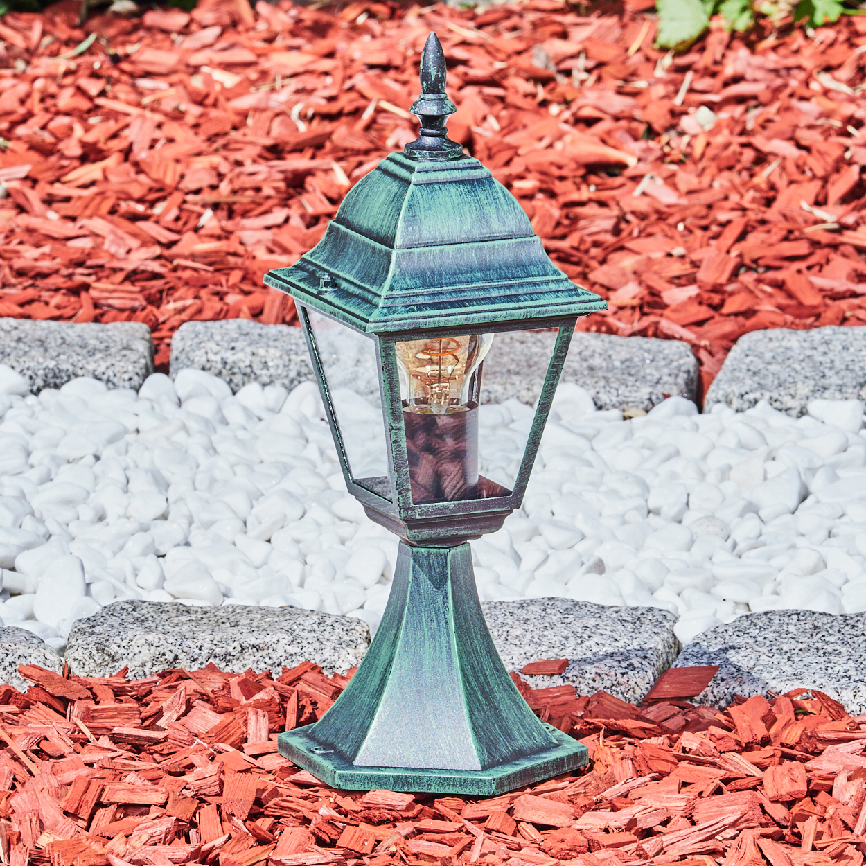 Eine grüne Lampe mit klarem Glas, auf roter Mulch, weißen Steinen und einem grünen Hintergrund.