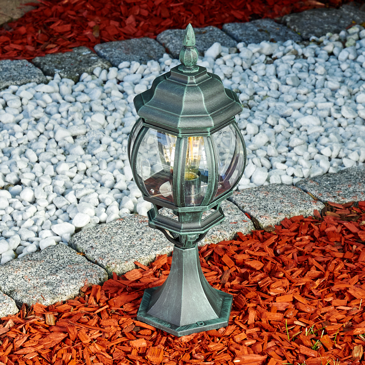 Grün-schwarze Außenlampe. Sie steht auf roter Mulch, weißen Steinen und grauen Steinziegeln.