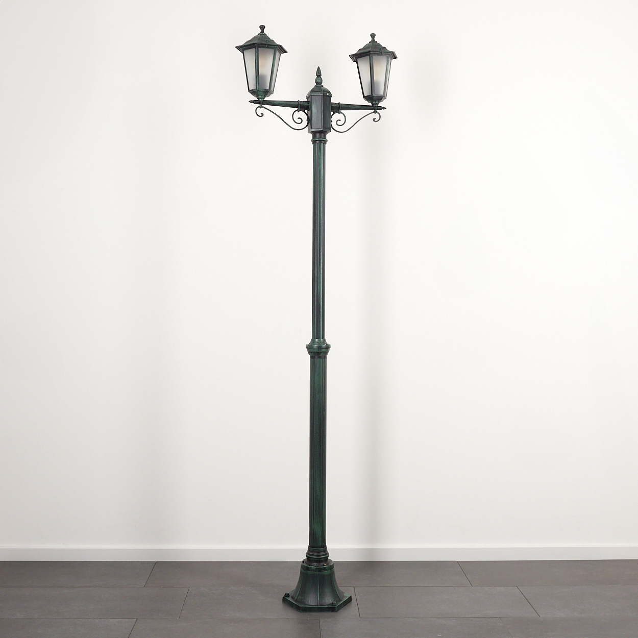 Eine hohe, grün-schwarze Straßenlaterne mit zwei Lampen. Sie steht auf einem grauen Steinboden vor einer weißen Wand.