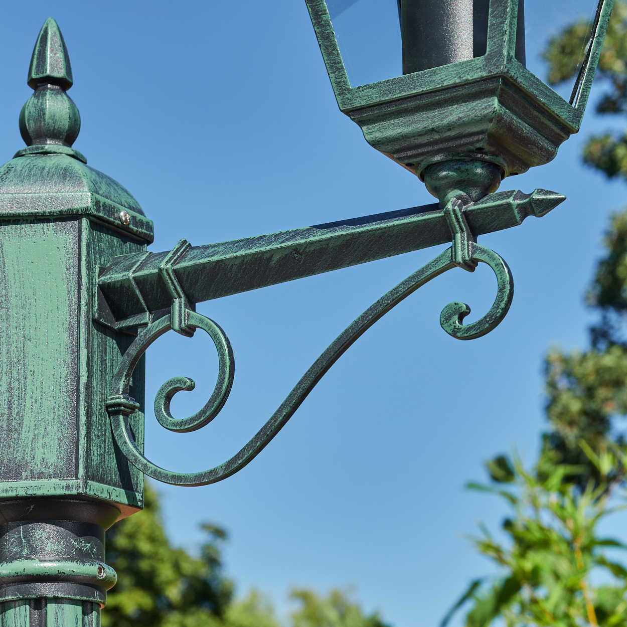Nahaufnahme des dekorativen Arms einer grünen Laterne. Es hat ein geschwungenes Design. Der Himmel ist blau.