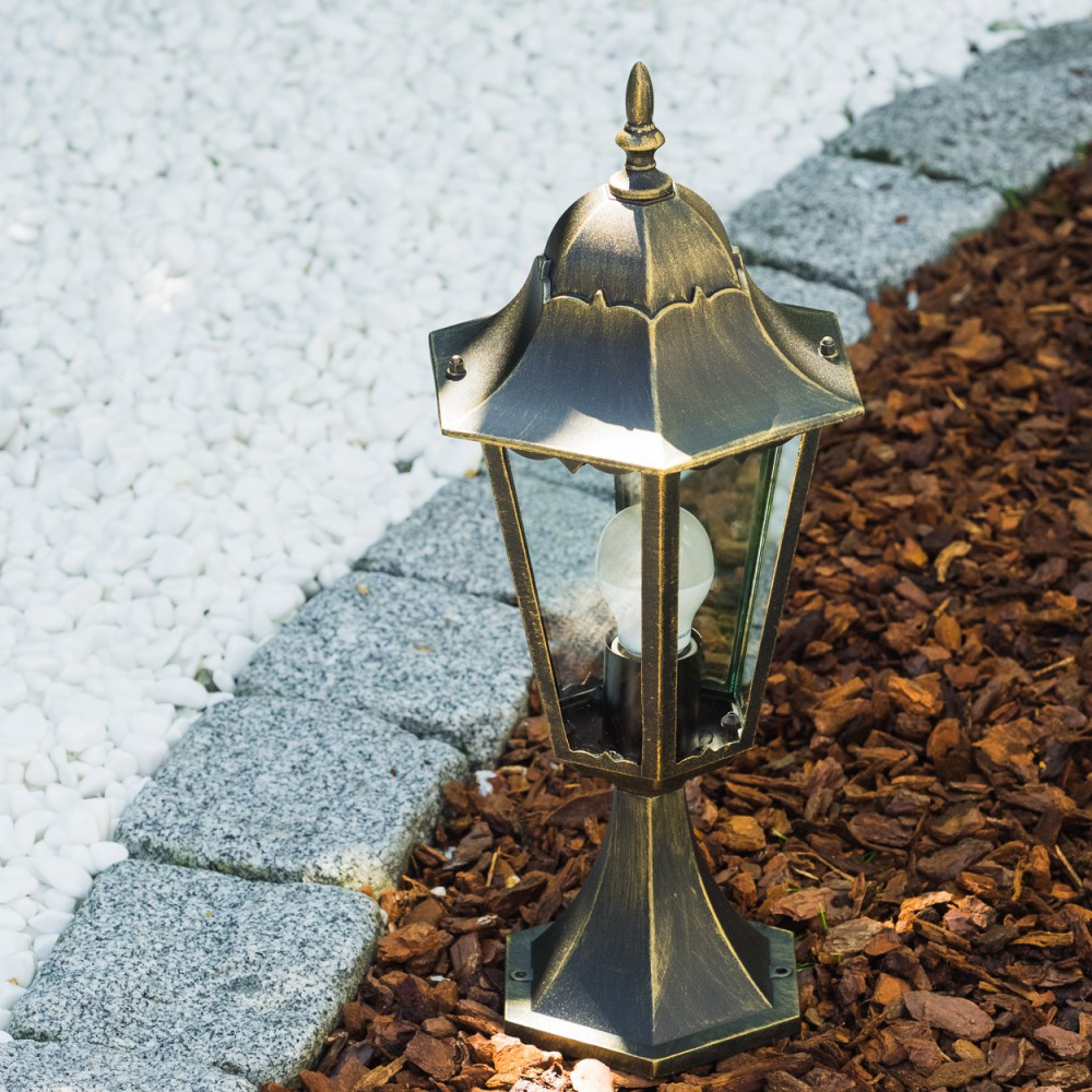 Außenlampe. Die Lampe ist golden mit klarem Glas und dekorativer Spitze. Garten, weiße Steine und rotes Mulch.