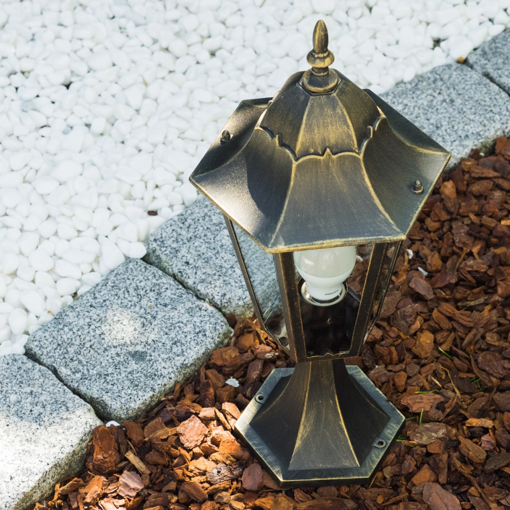 Eine bronzefarbene Lampe mit weißer Glühbirne steht auf braunem Mulch, begrenzt von grauen Steinen und weißen Kieselsteinen.
