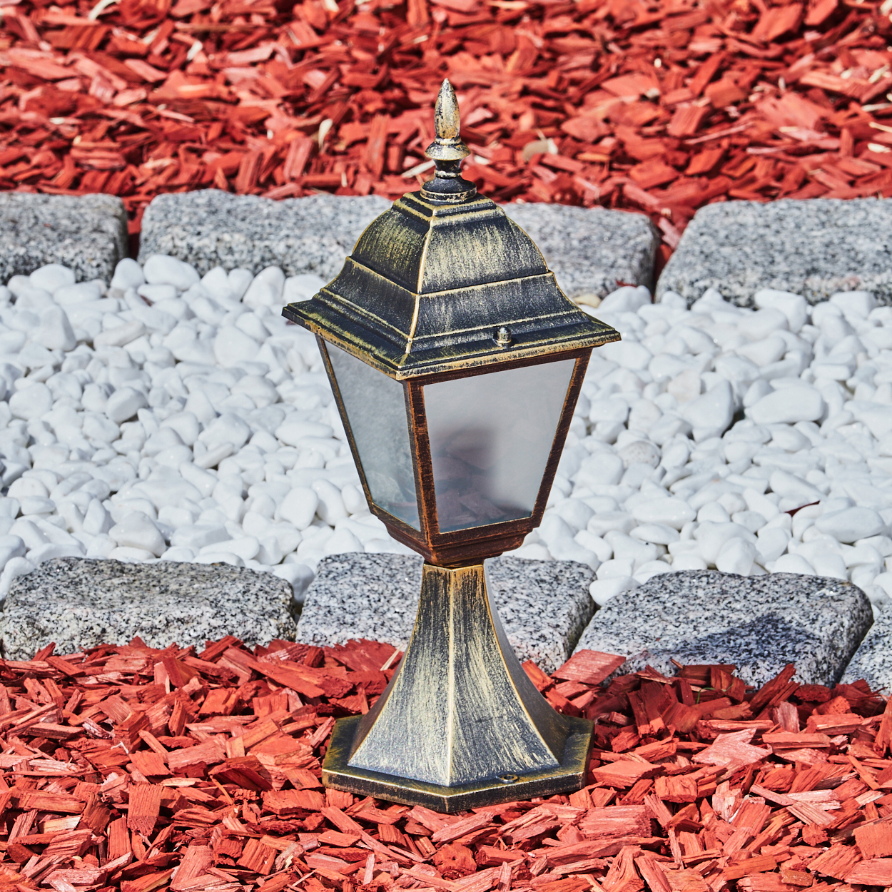 Goldene Außenlampe auf roter Mulch, weißen Steinen. Die Lampe hat klares Glas und eine dekorative Spitze. Garten.