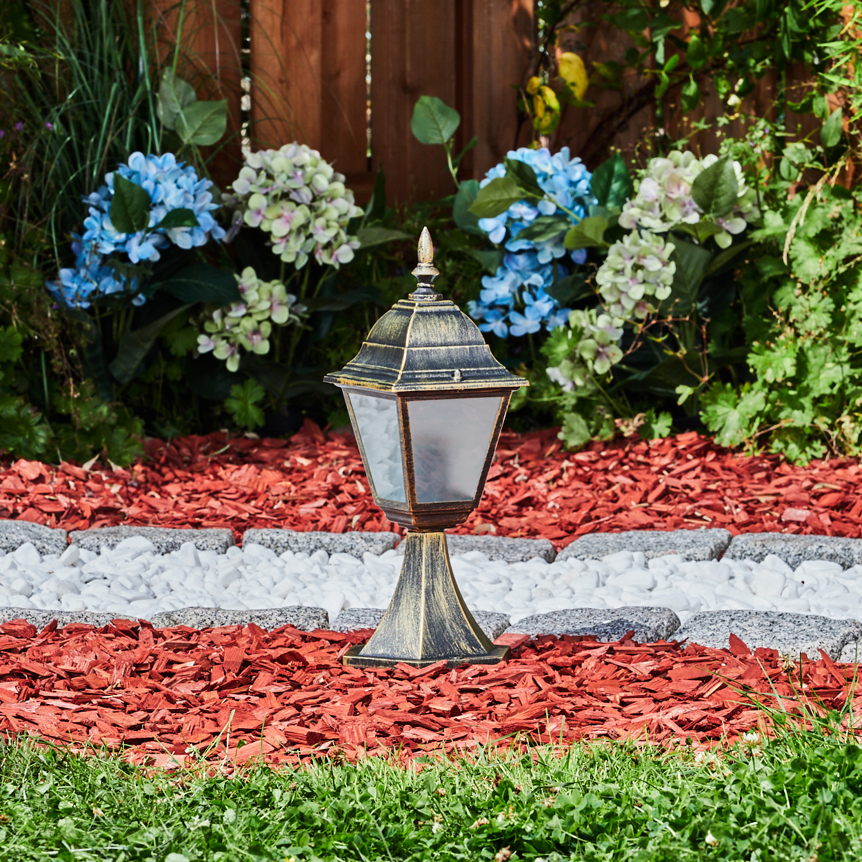 Außenlampe in einem Garten. Die Lampe ist golden mit klarem Glas und dekorativer Spitze. Umgeben von Pflanzen.