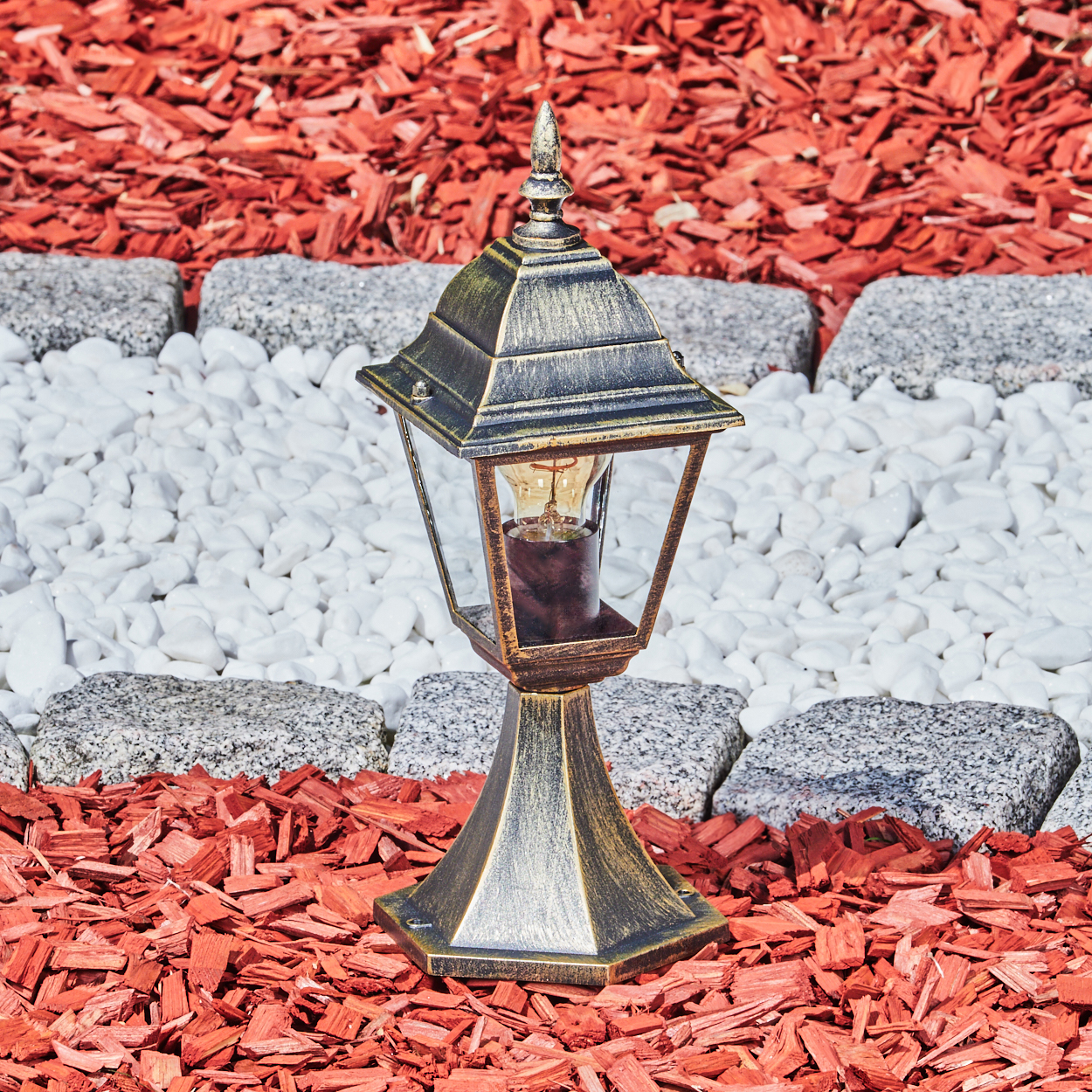 Eine Außenlampe mit goldfarbenem Finish auf roter Mulch, weißen Steinen und einem Steinrand.