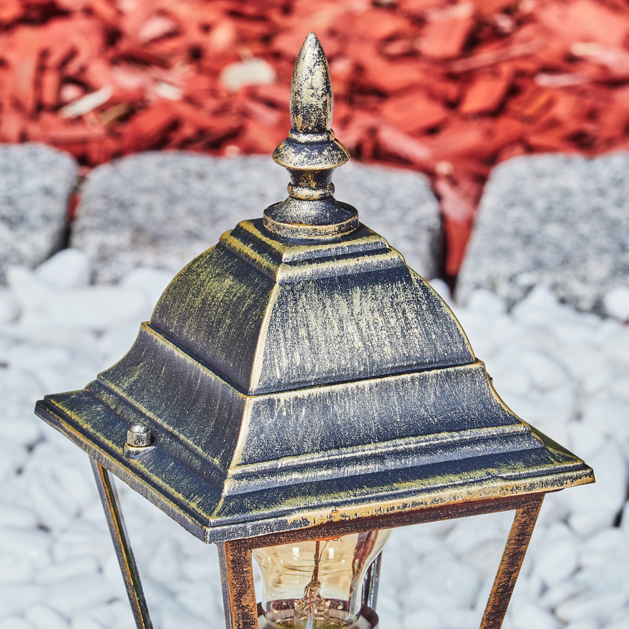Nahaufnahme einer Außenlampe mit goldfarbenem Finish auf roter Mulch und weißen Steinen, mit leuchtender Birne.
