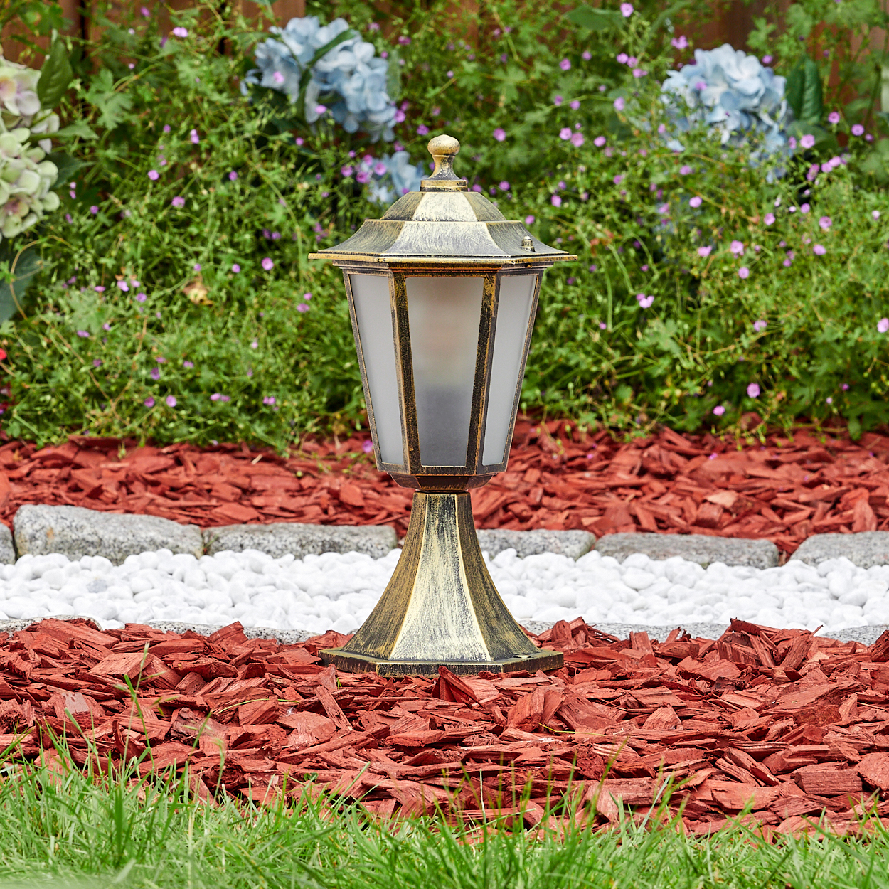 Außenlampe mit goldfarbenem Finish, umgeben von roter Mulch, weißen Steinen und Grün, mit Blumen im Hintergrund.