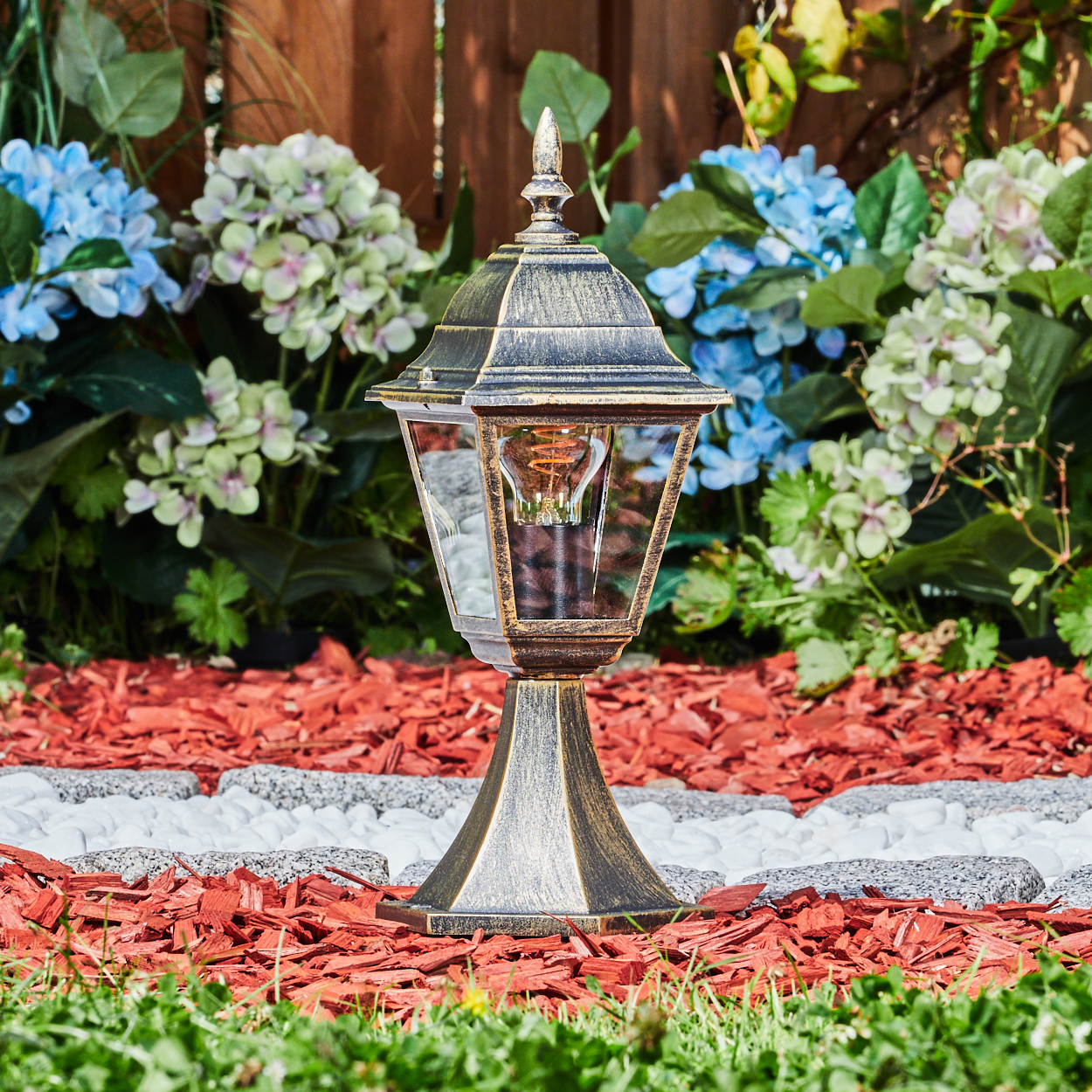 Bronze-Lampe im Garten, Glühbirne in Glaseinfassung, umgeben von Blumen.