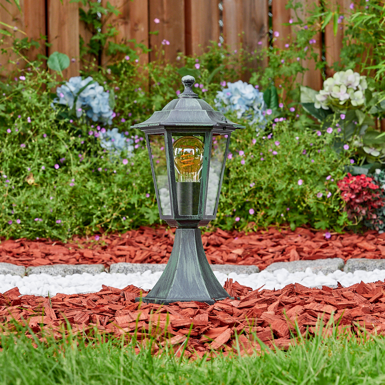 Bronze-Lampe im Garten, Glühbirne, auf roter Mulch, umgeben von Grün.