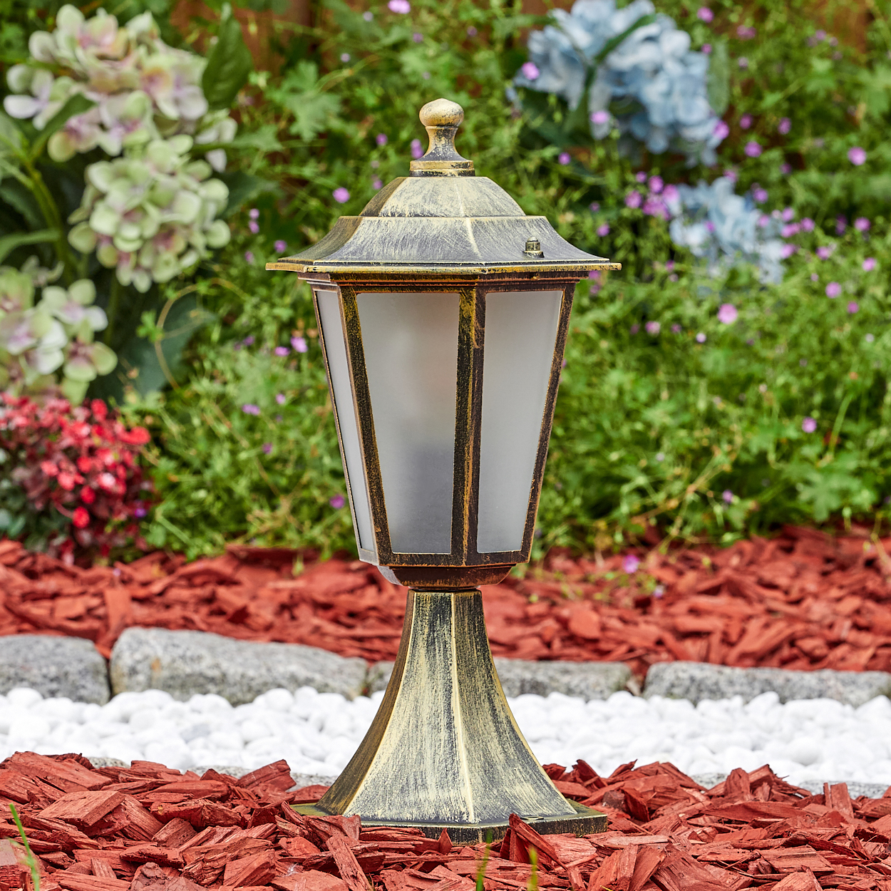Bronze-Lampe im Garten, Milchglas, auf roter Mulch, umgeben von Blumen.
