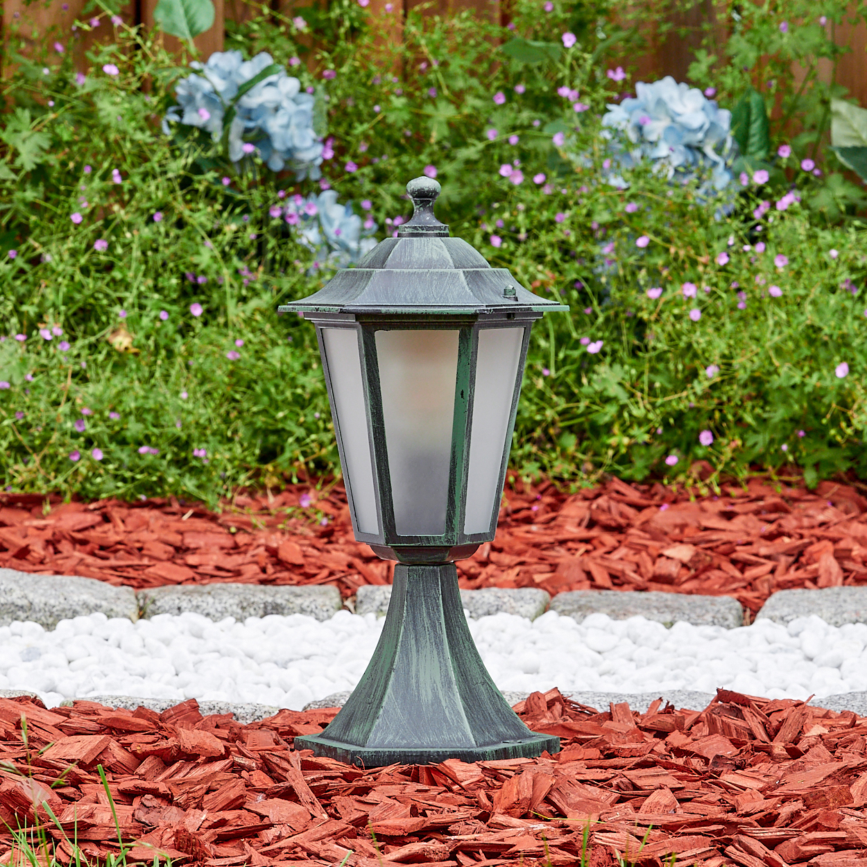 Außenlampe in einem Garten mit roter Mulch, weißen Steinen und blauen Blumen.