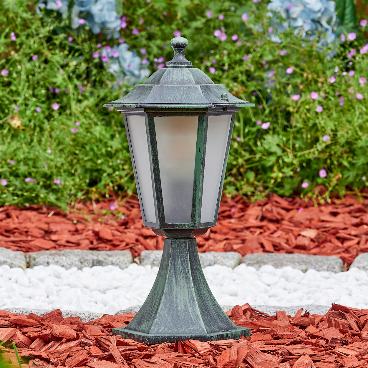 Eine grüne Außenlampe steht auf roter Mulche. Blumen sind im Hintergrund.