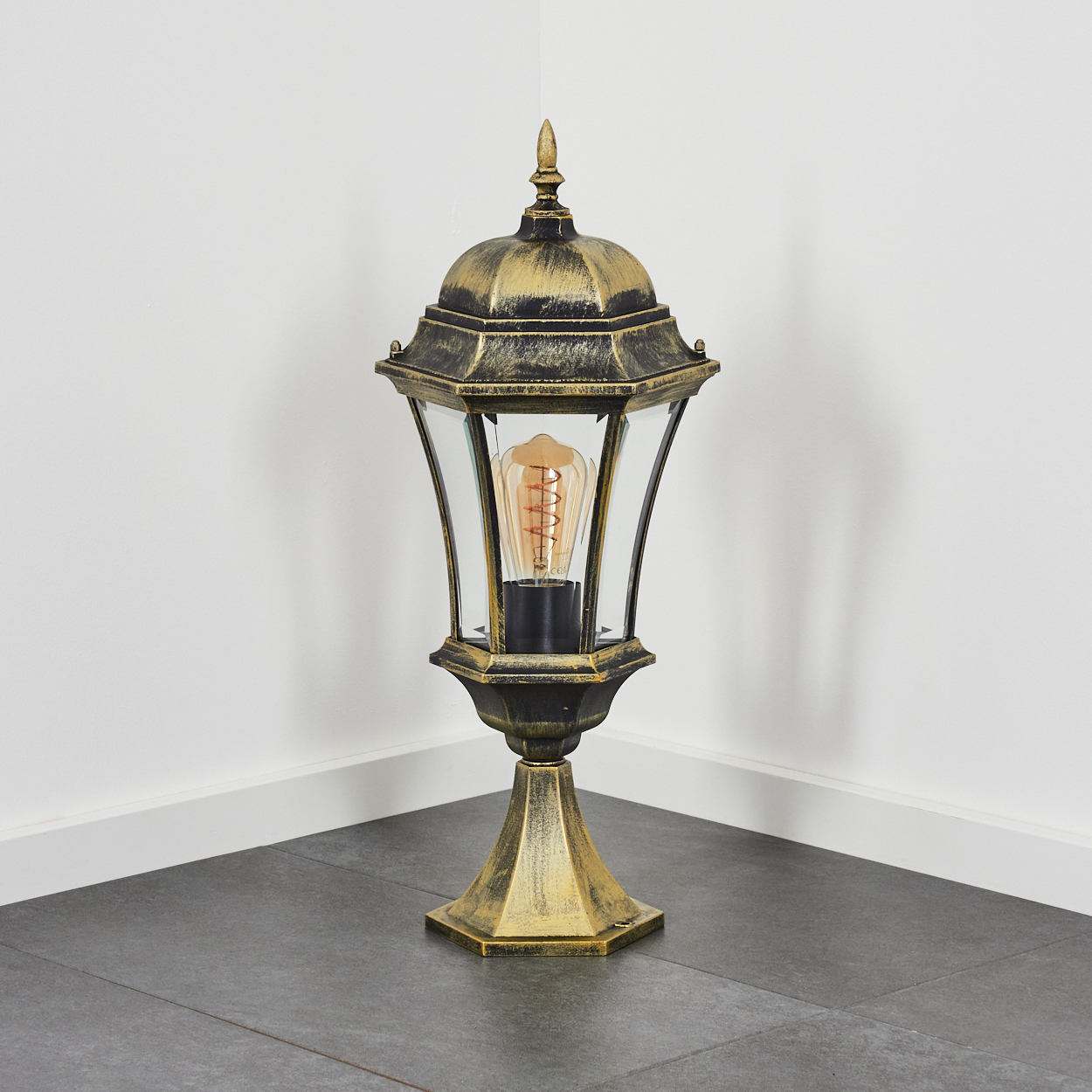 Goldene und schwarze Außenlampe in einer Ecke. Die Lampe hat eine Glühbirne. Der Boden besteht aus grauen Fliesen.