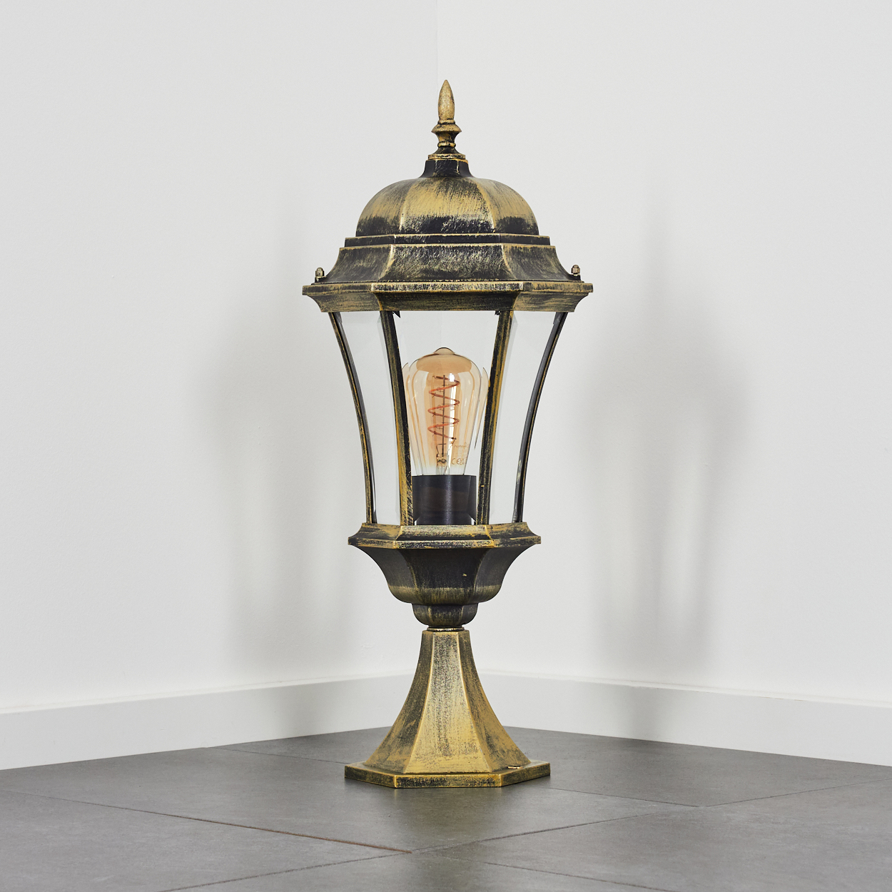 Goldene und schwarze Außenlampe auf dunklem Boden in der Ecke eines Raumes. Weiße Wände im Hintergrund.