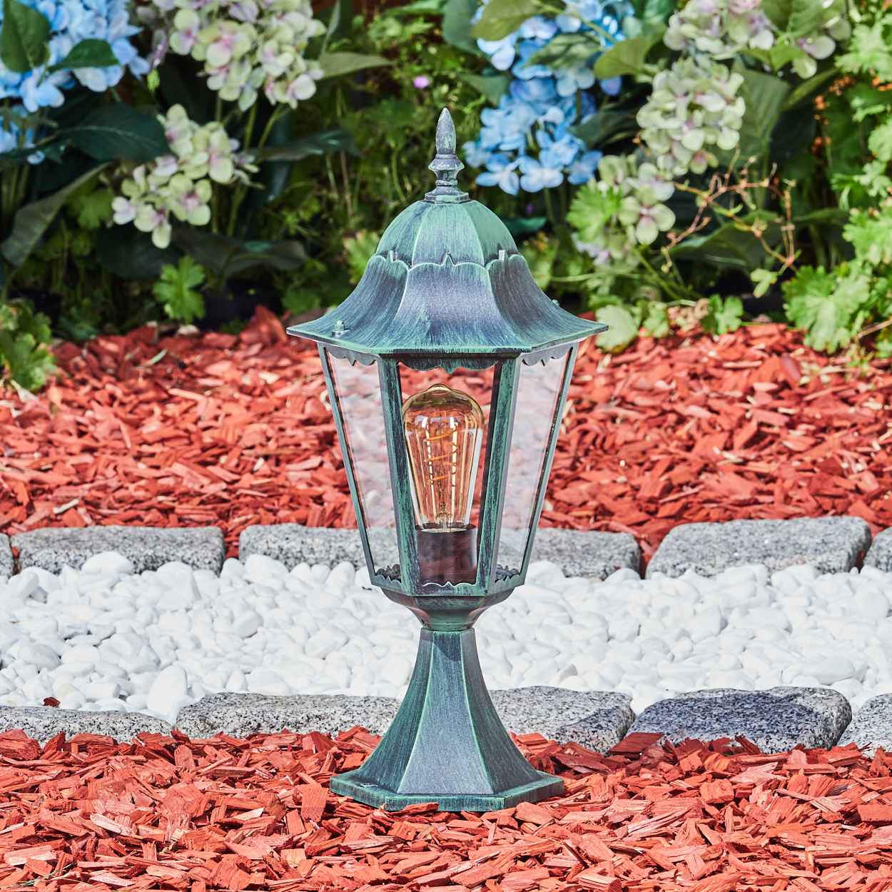 Grüne Außenlampe mit Glühbirne im Gartenbeet mit roter Mulch, weißen Steinen und Blumen im Hintergrund.