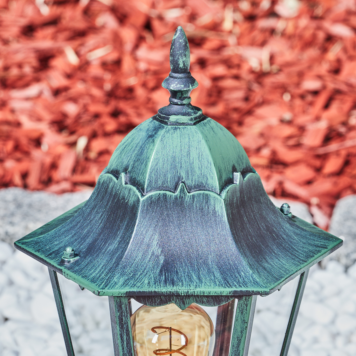 Nahaufnahme der Oberseite einer grünen Außenlampe mit Glühbirne. Roter Mulch und weiße Steine im Hintergrund.