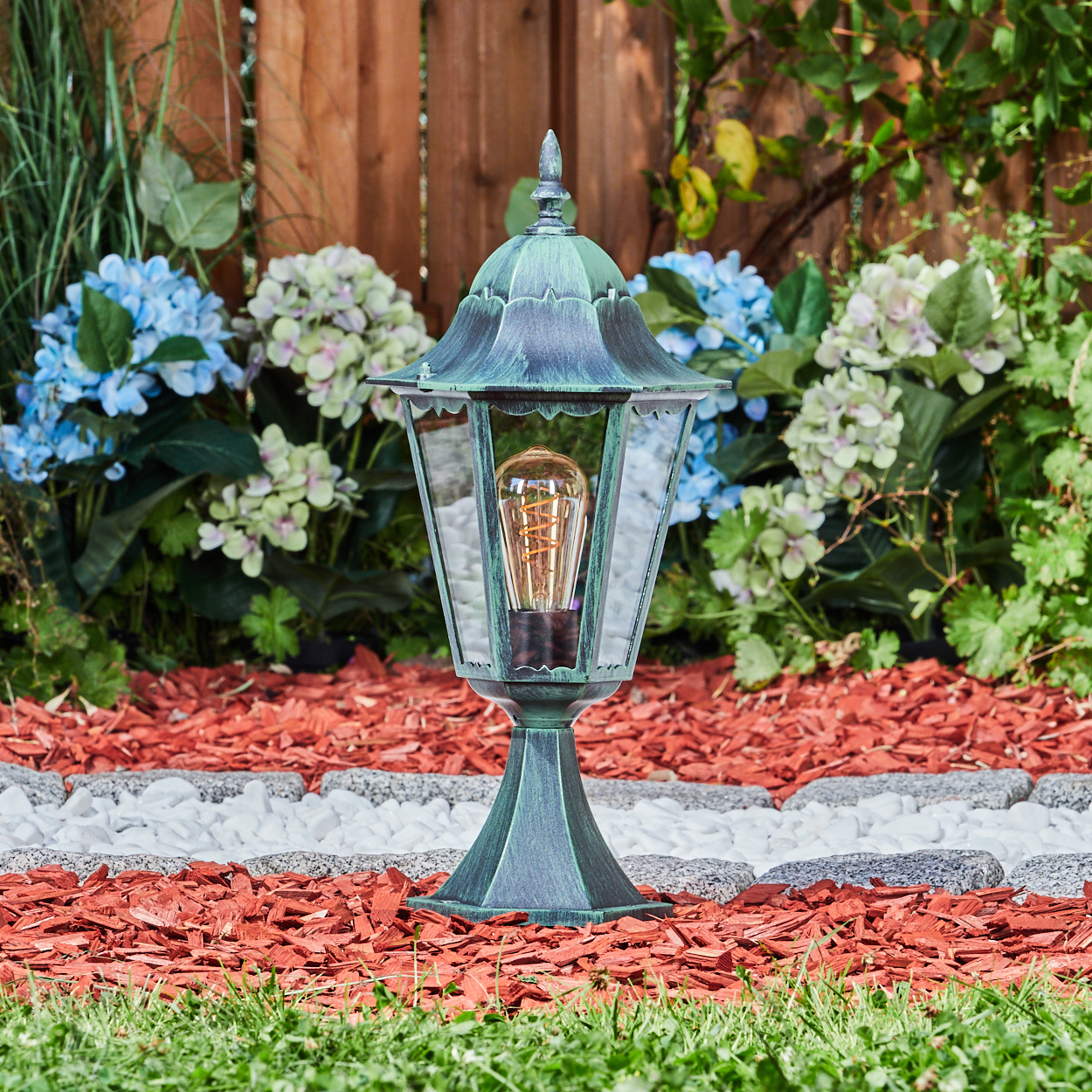 Grüne Außenlampe in einem Garten. Roter Mulch, weiße Steine und Gras im Vordergrund. Blaue Blumen im Hintergrund.