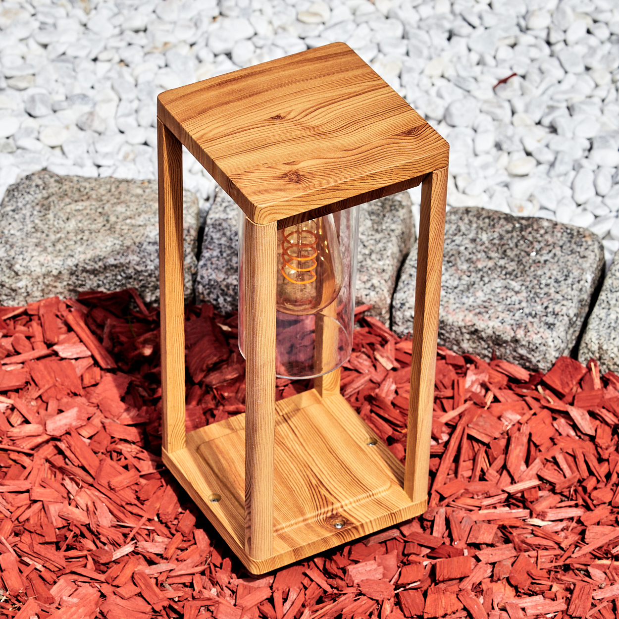 Holzlampe mit einer Glühbirne in einem Glasrohr. Roter Mulch und weiße Steine im Hintergrund.