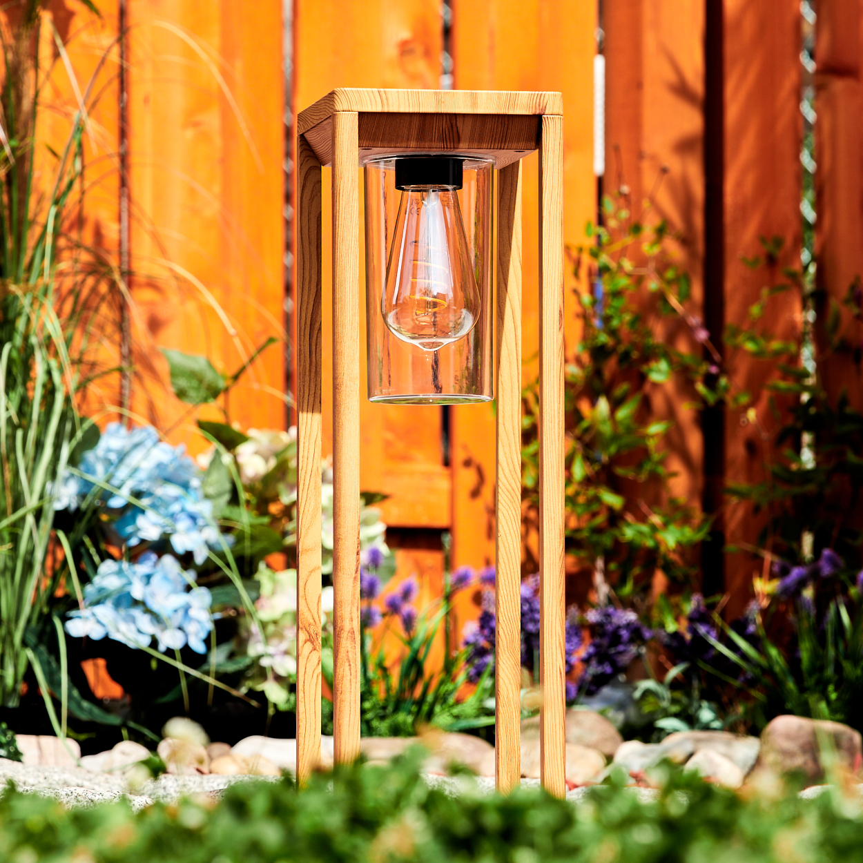 Holz-Außenlampe mit Glühbirne in einem Glaszylinder. In einem Garten mit Blumen und Holzzaun aufgestellt.