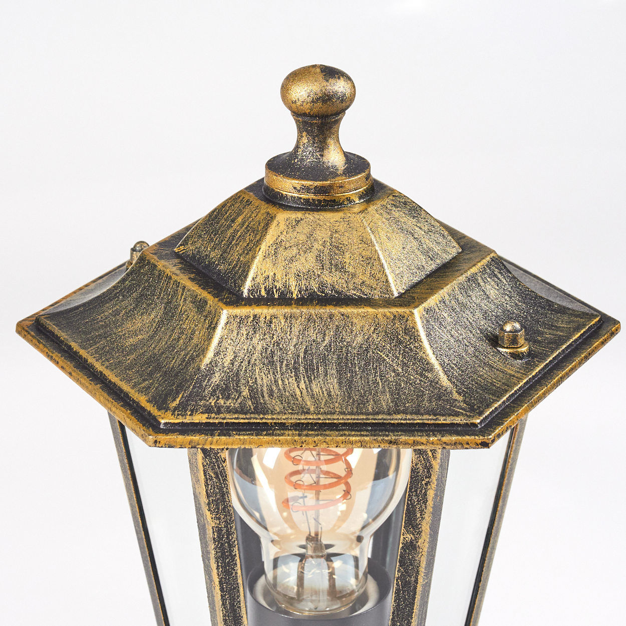 Nahaufnahme einer dekorativen Außenlampe mit gold-schwarzem Finish. Die Lampe hat ein Glasgehäuse und eine dekorative Spitze.