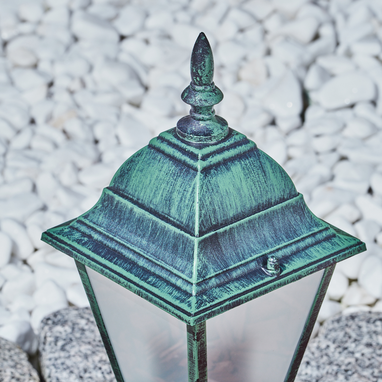 Eine grün-schwarze Außenlampe mit dekorativer Spitze, vor dem Hintergrund weißer Steine.