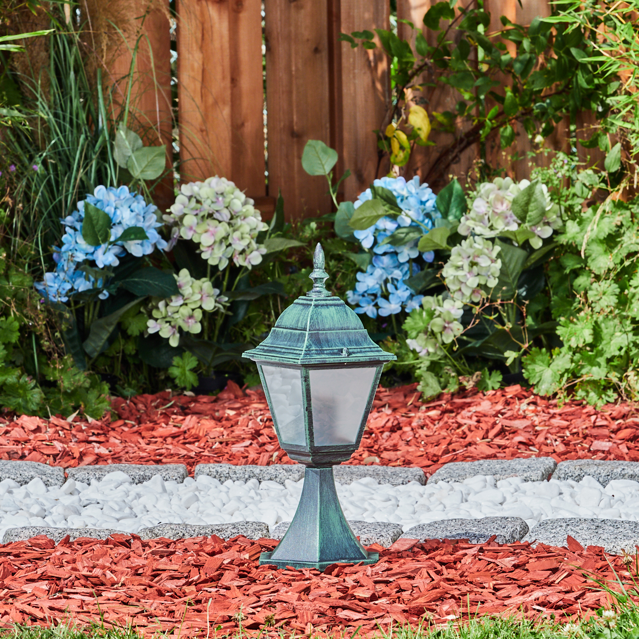 Eine Außenlampe mit grüner Oberfläche, in einem Garten mit Blumen, roter Mulch und weißen Steinen.
